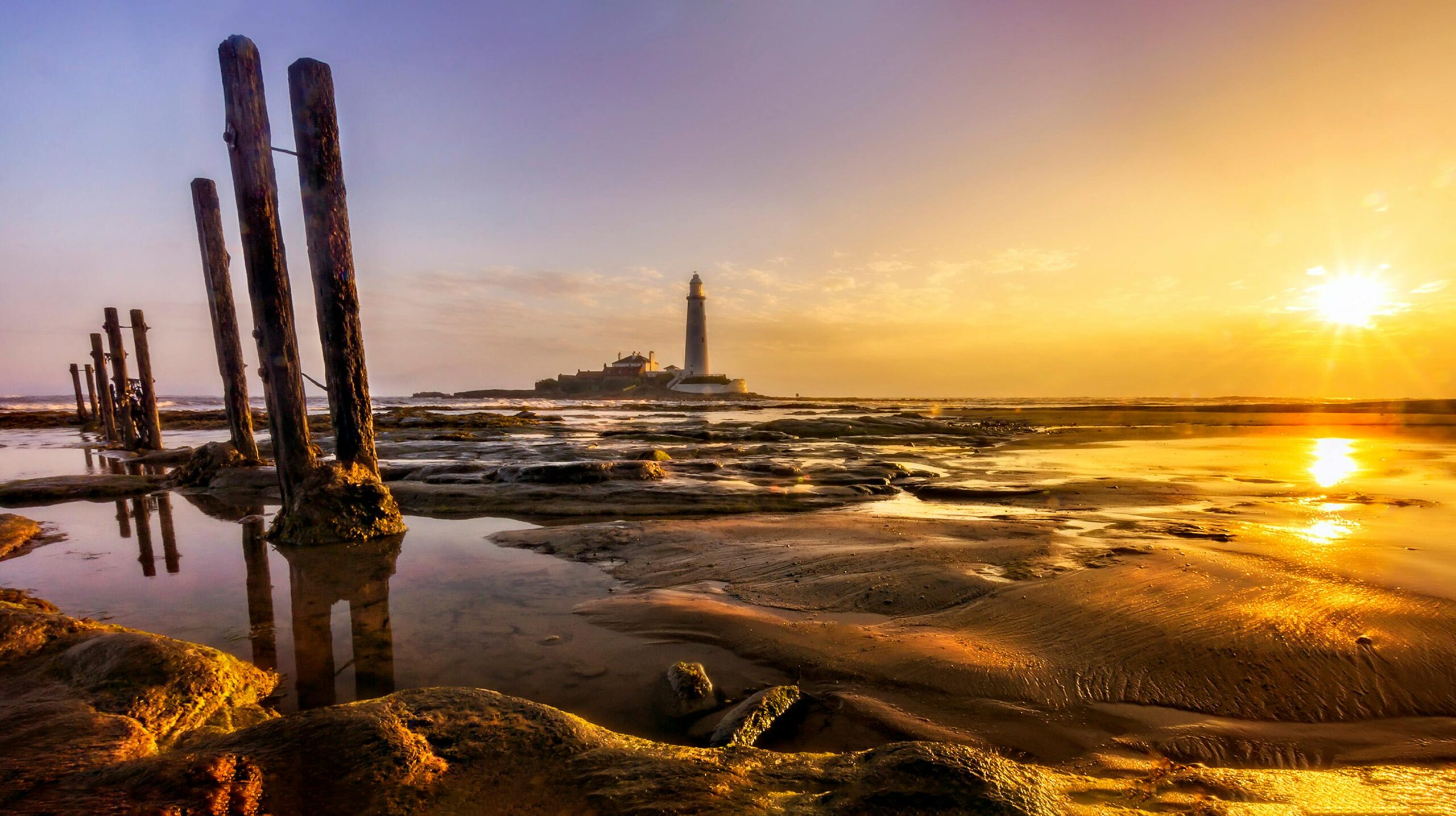 Golden sunset over a rocky shoreline with a lighthouse in the distance, tranquil scenery.